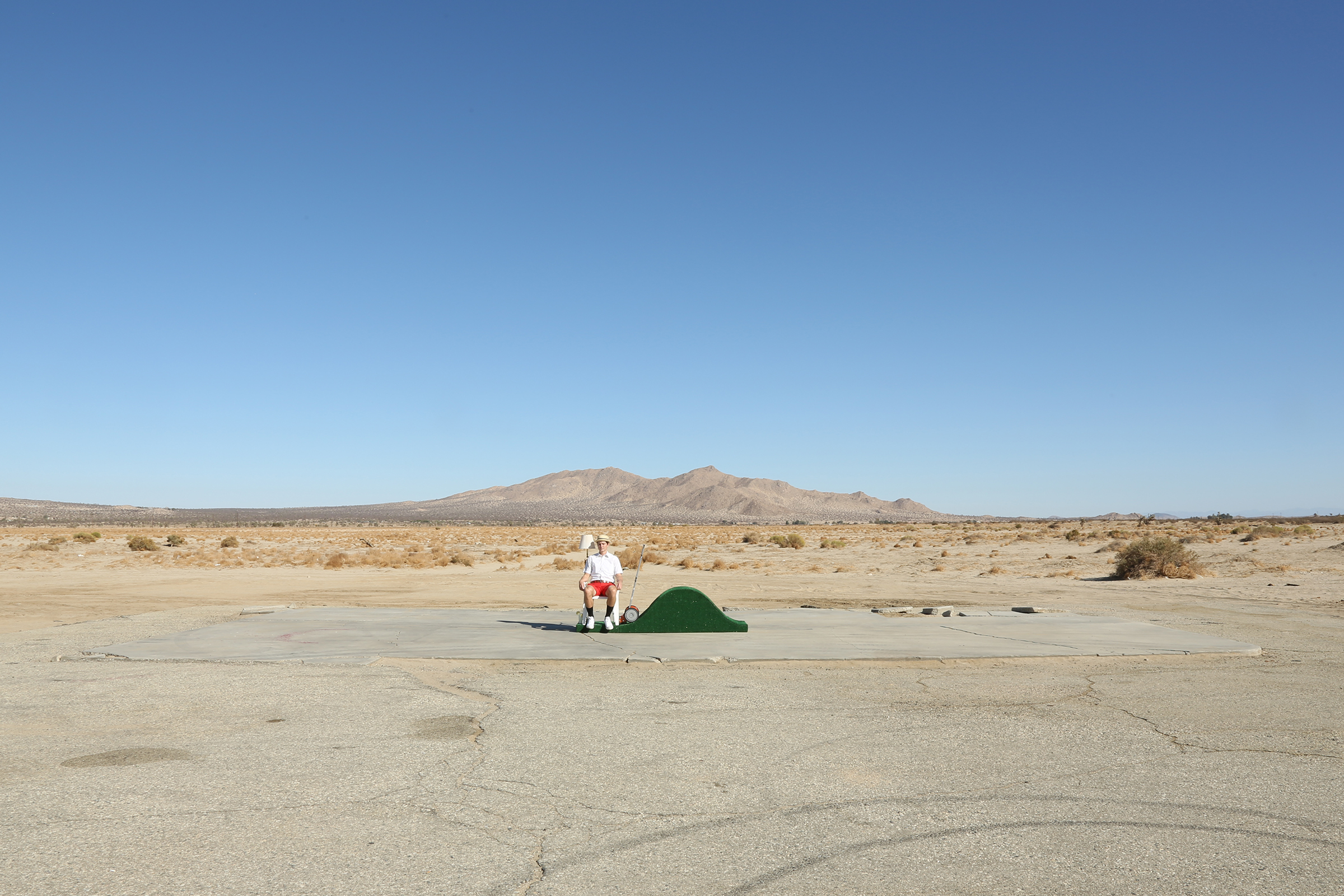 A person sits next to a small bell-curve shaped mound of artificial turf in the middle of desert.
