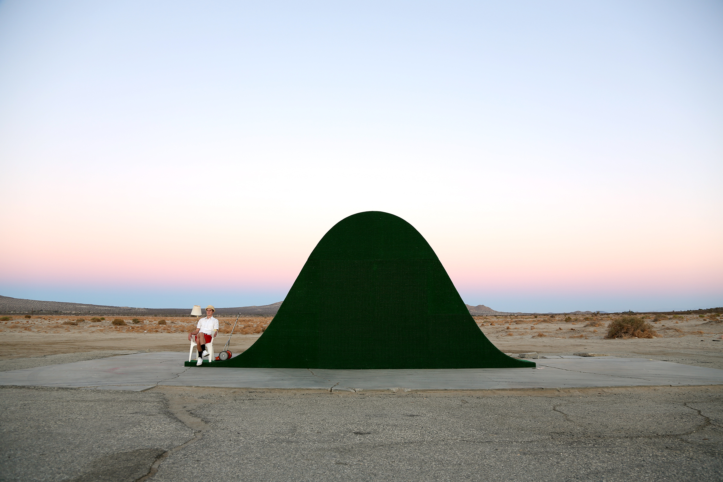 A person sits next to a large bell-curve shaped mound of artificial turf in the middle of desert.