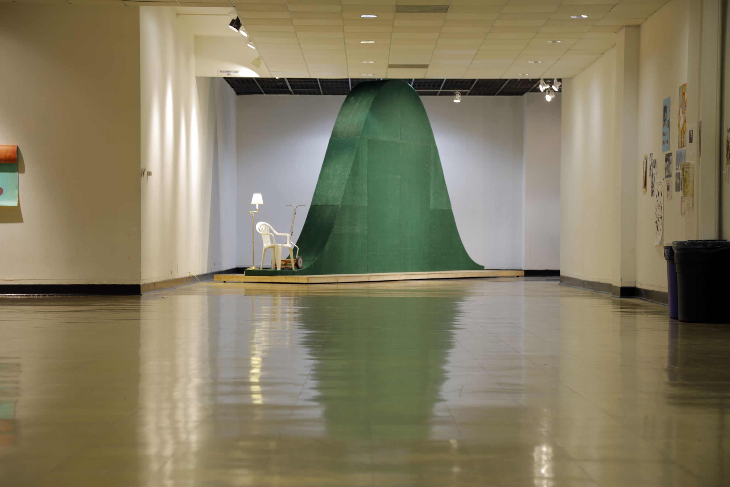 A photograph of a white plastic lawn chair next to a large, green, bell-curve shaped sculpture in a school hallway