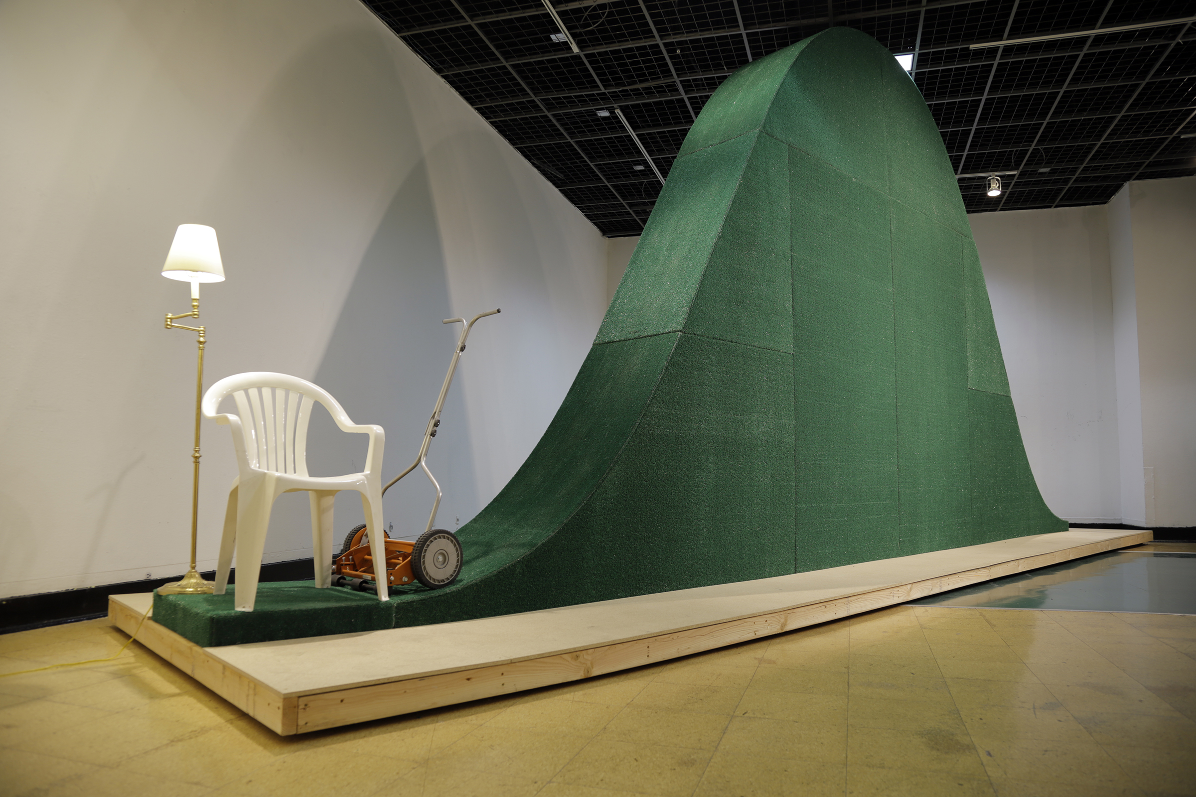 A close-up photograph of white plastic lawn chair, floor lamp, and push reel mower next to a large, green, bell-curve shaped sculpture.
