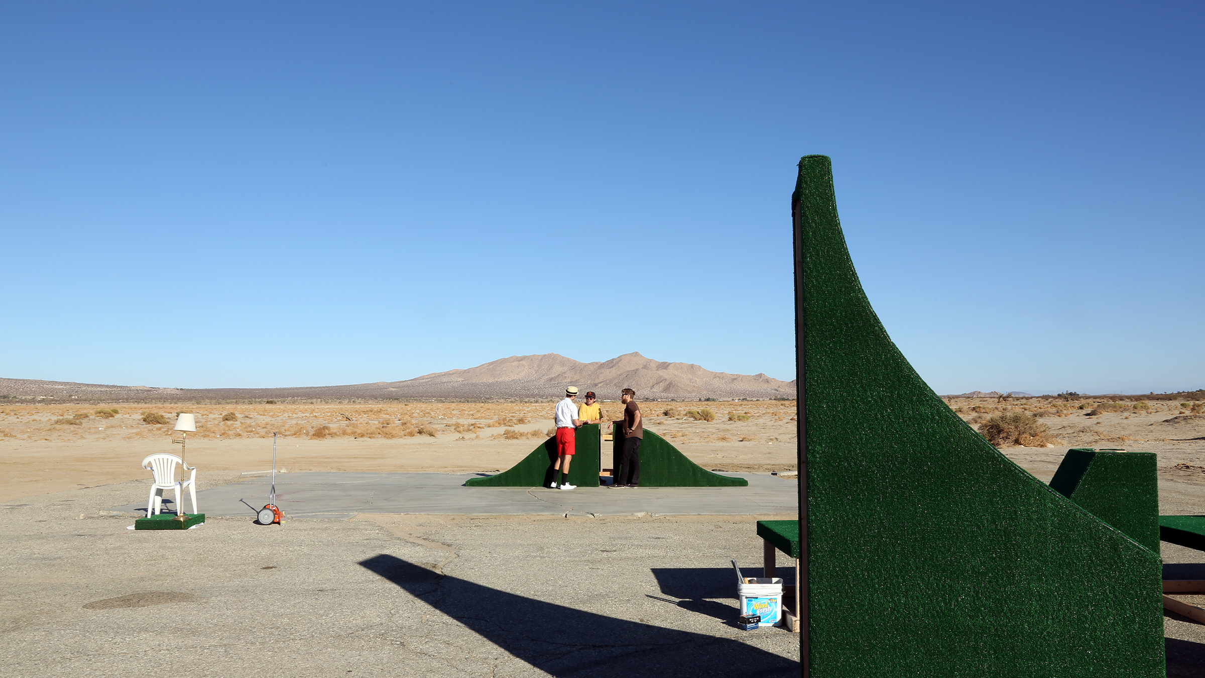 Three people chatting next to green, curved sculptures in the middle of desert shrubland.