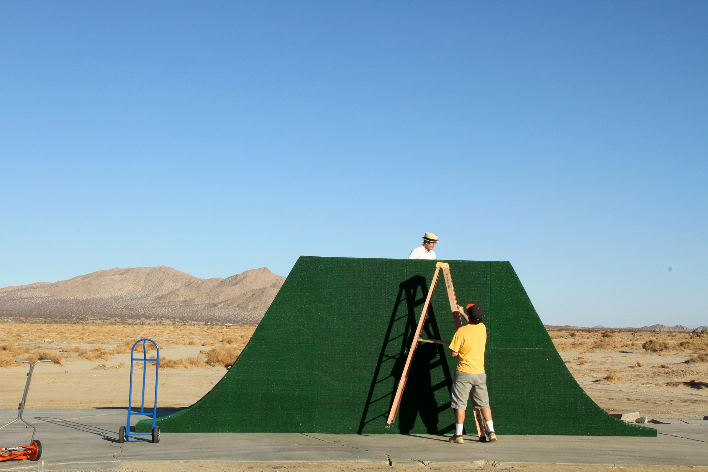 Two people assembling pieces of a large green sculpture in the middle of desert shrubland.