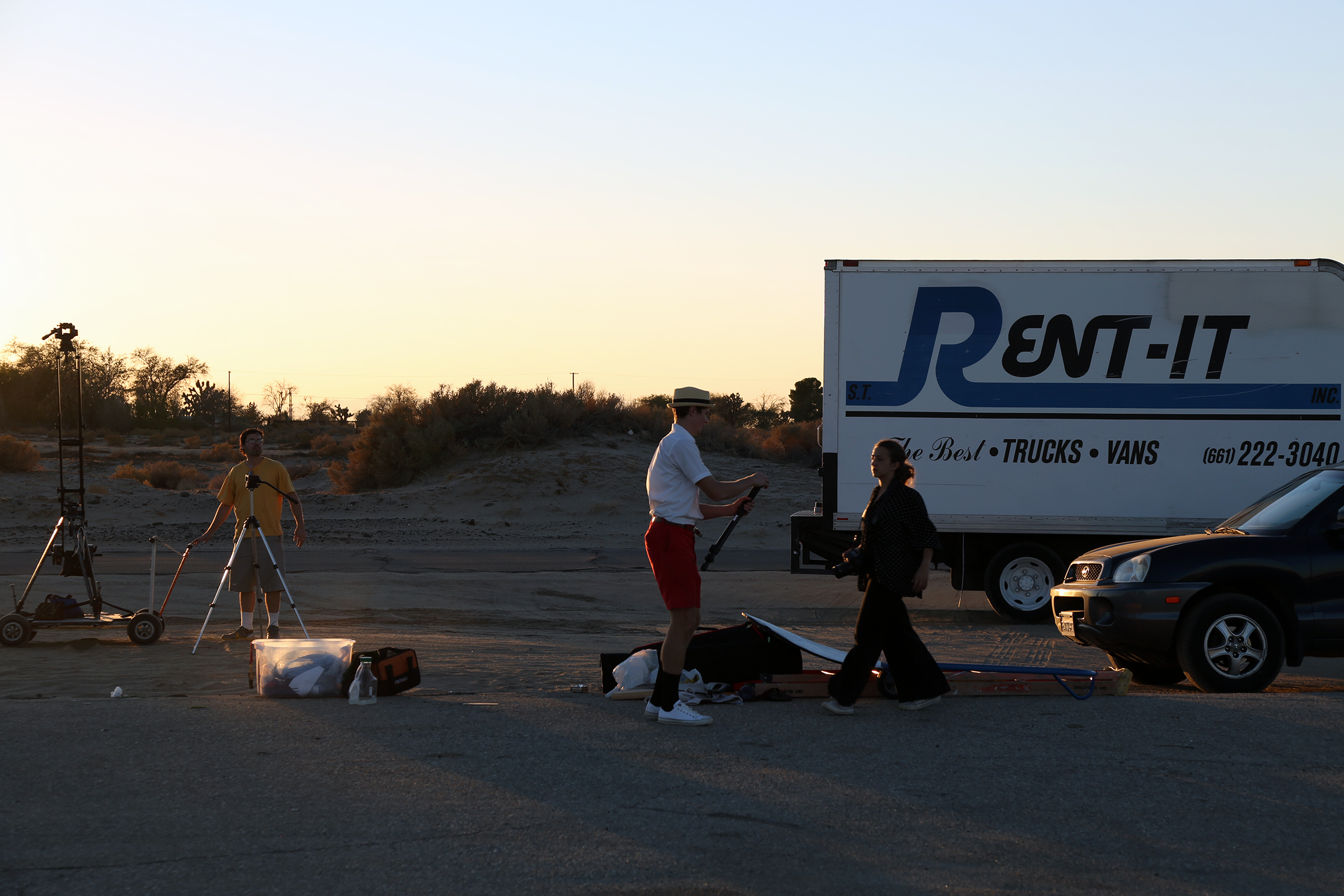Three people organizing filming equipment next to a box truck and car as the sun sets in the desert.