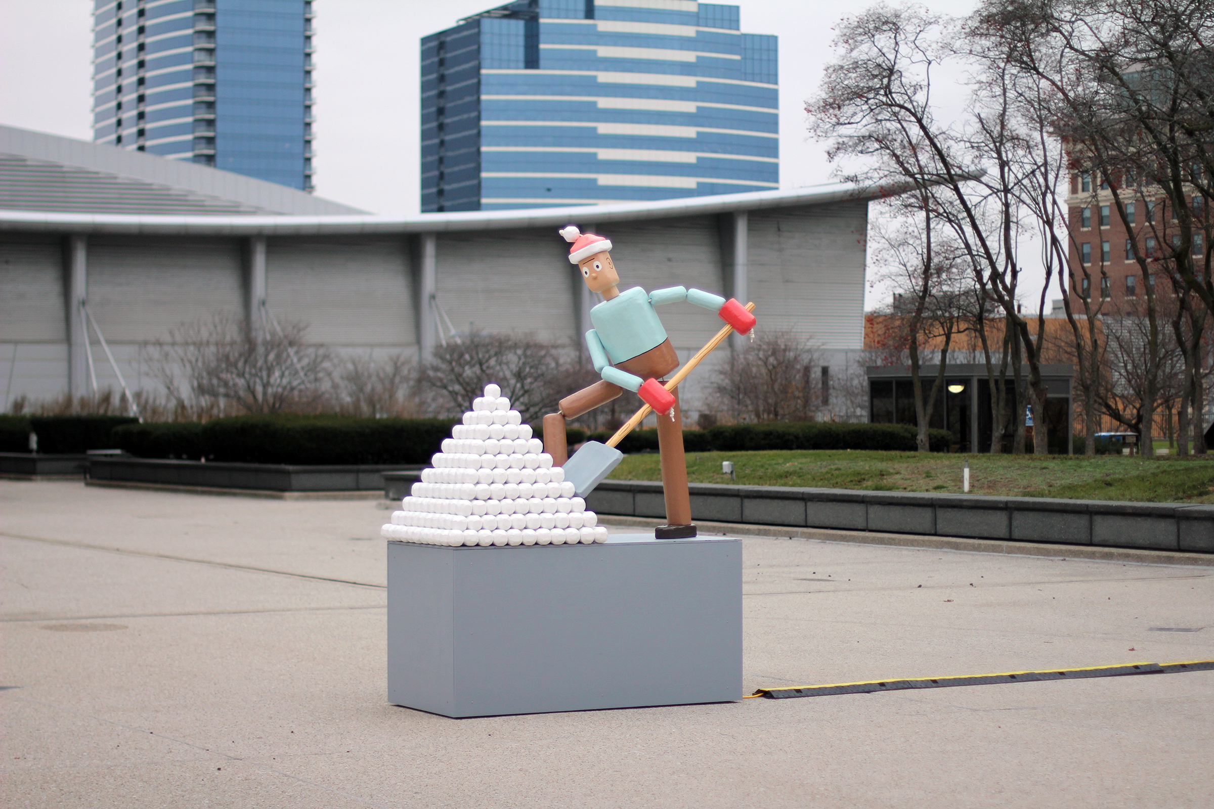 a photograph of a human-sized sculpture that looks like push puppet shoveling snow on a plinth in a concrete plaza