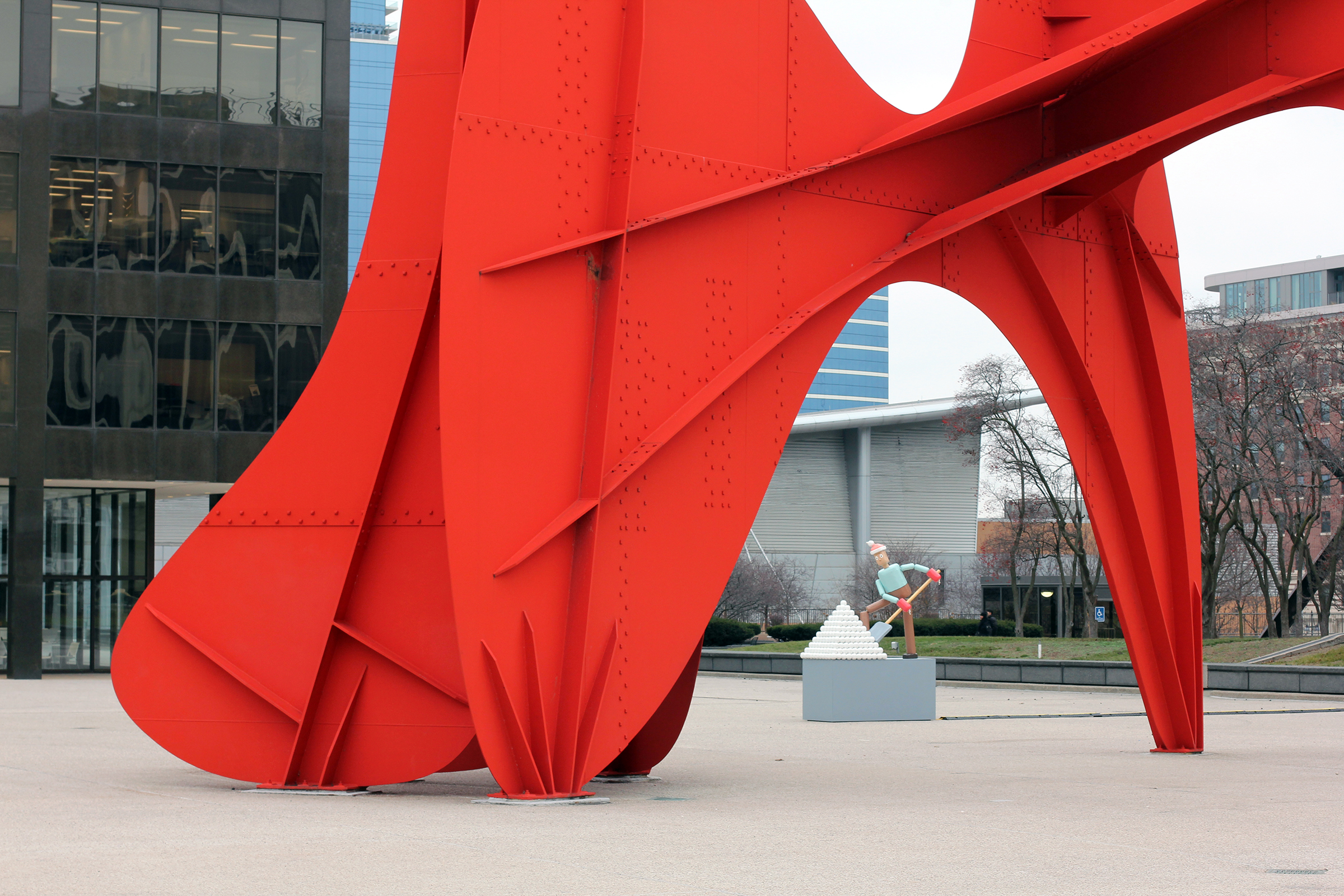 a life-size push puppet sculpture in a concrete plaza with a two-story red, metal, abstract sculpture in the foreground