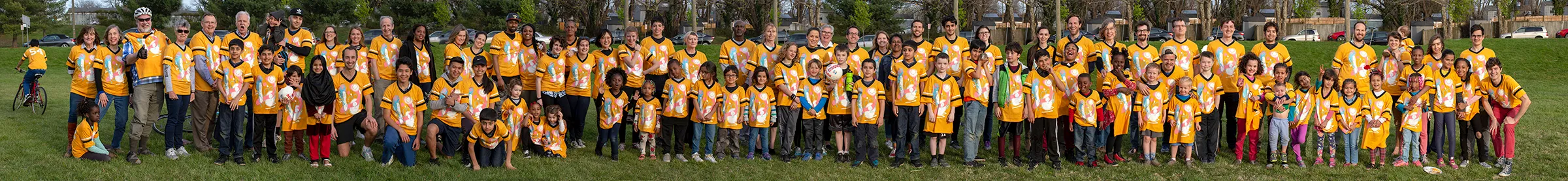 A extra-long horizontal photo of dozens of people wearing yellow soccer jerseys and standing in two rows in a grassy field.