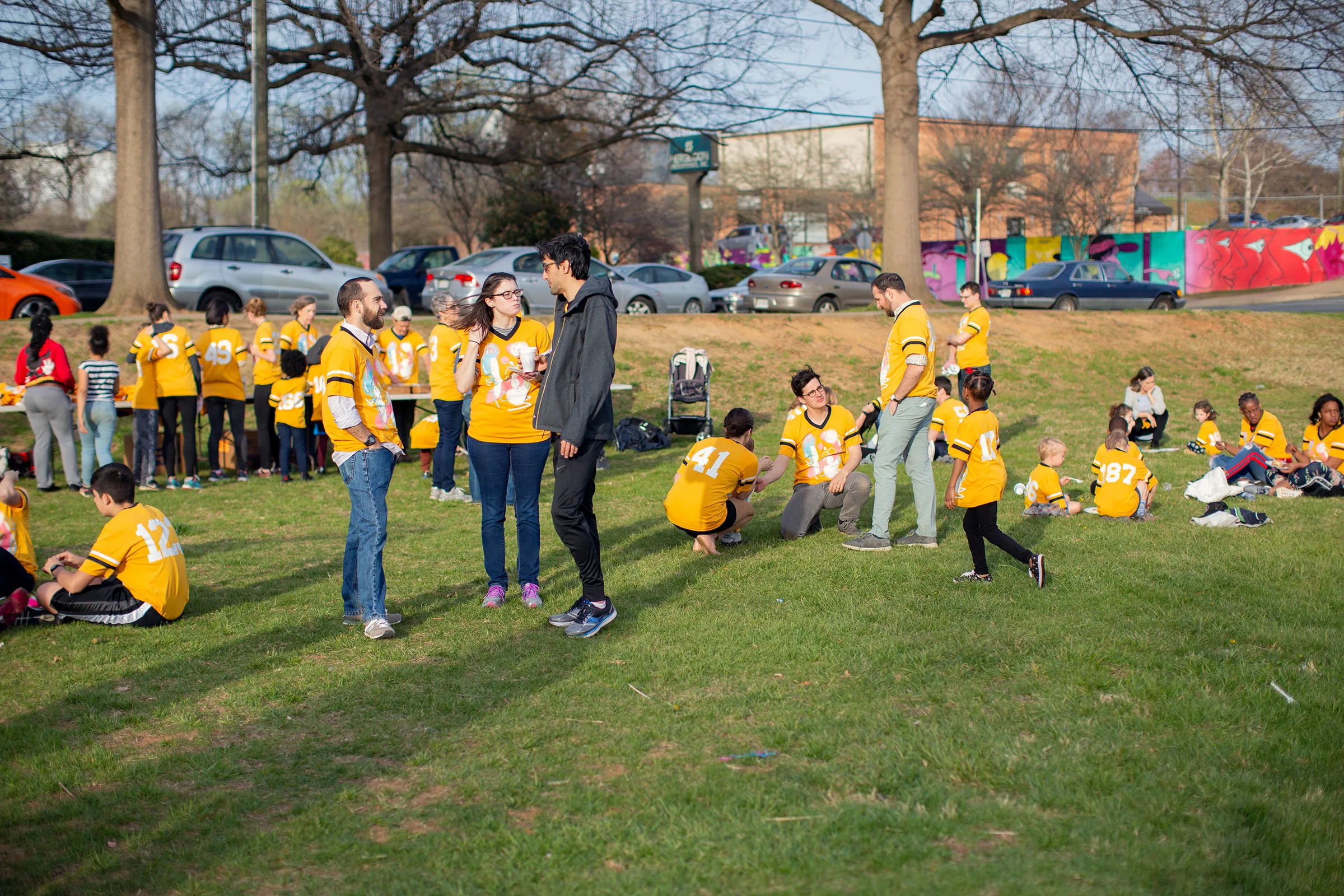 A photograph of a group of two dozen people chatting and eating a picnic dinner on a grass field. Most wear matching yellow jerseys.