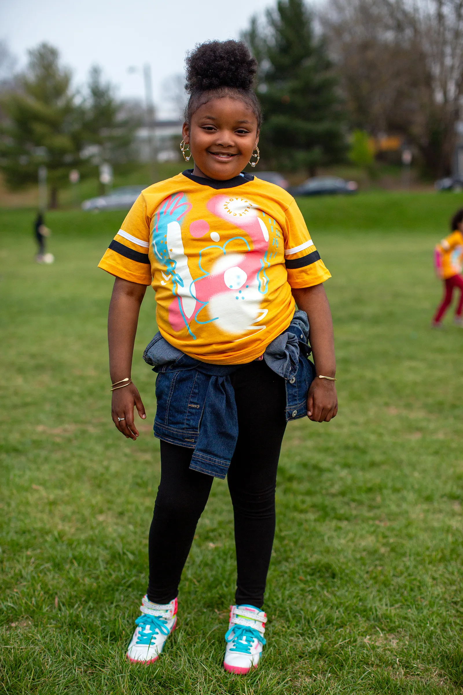 A photograph of a kid smiling while wearing a yellow soccer jersey with an abstract floral graphic printed on it. She has a jean jacket tied around her waist
