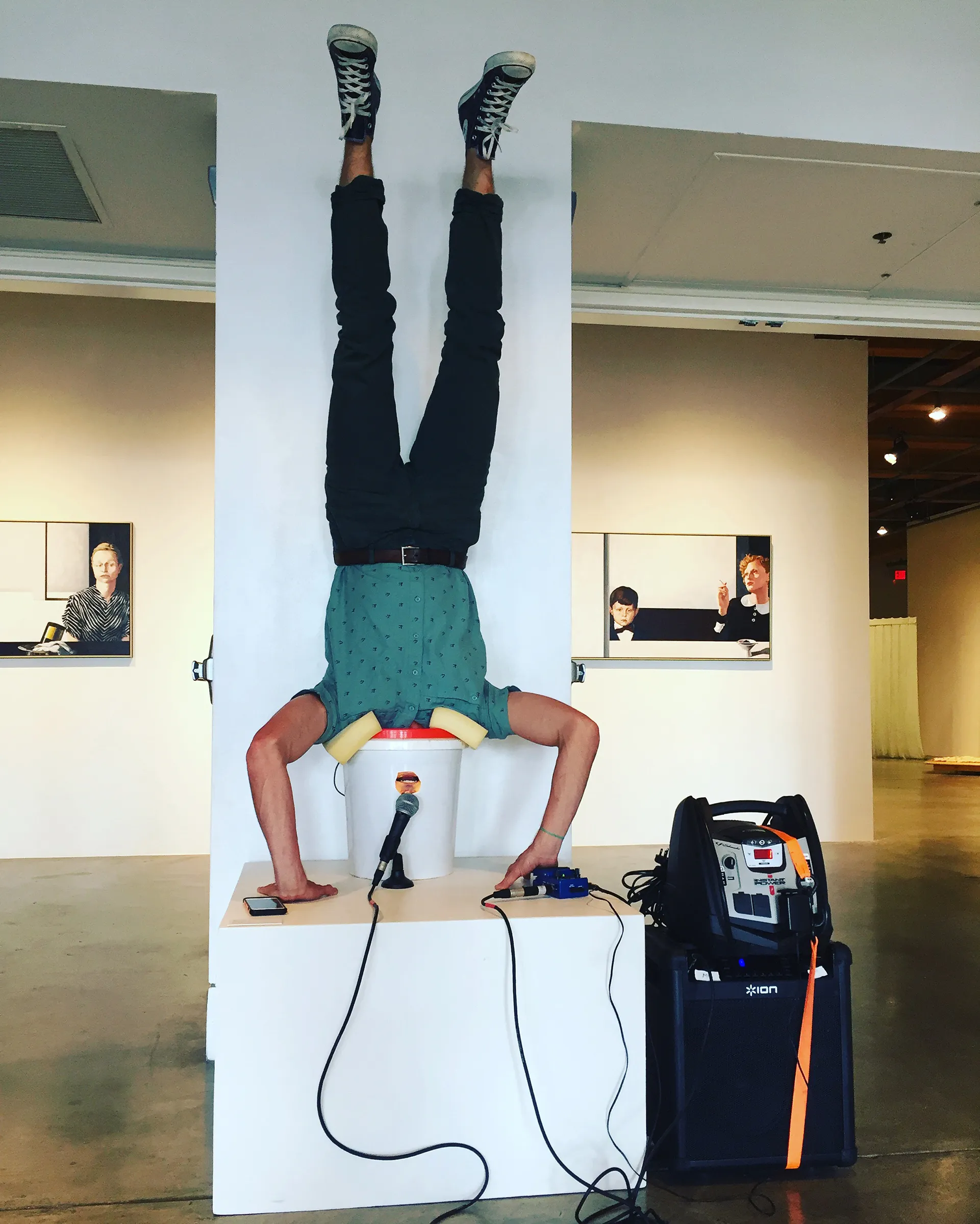 A photograph of Avery Lawrence doing a handstand with his head in a white 5-gallon bucket. He wears a green, short-sleeve, button-up shirt, black pants, and gray high top Converse All-Stars.