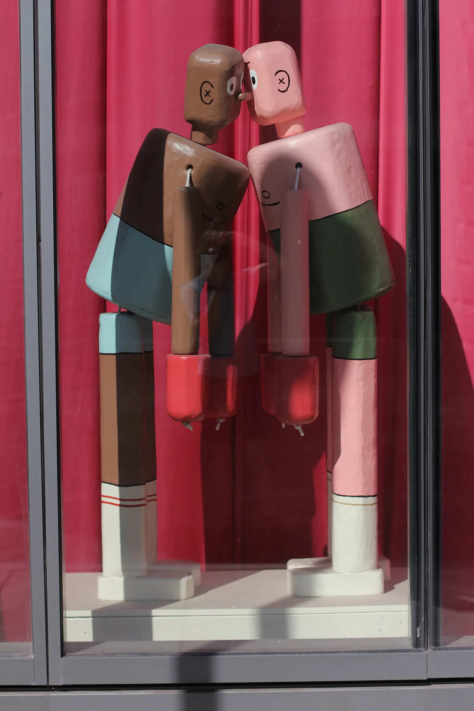 A photograph of Boxers. Two life-sized push puppets are painted to look like boxers. They have collapsed into each other, cheek to cheek, chest to chest, arms pulled straight down by the weight of chunky, red boxing gloves.