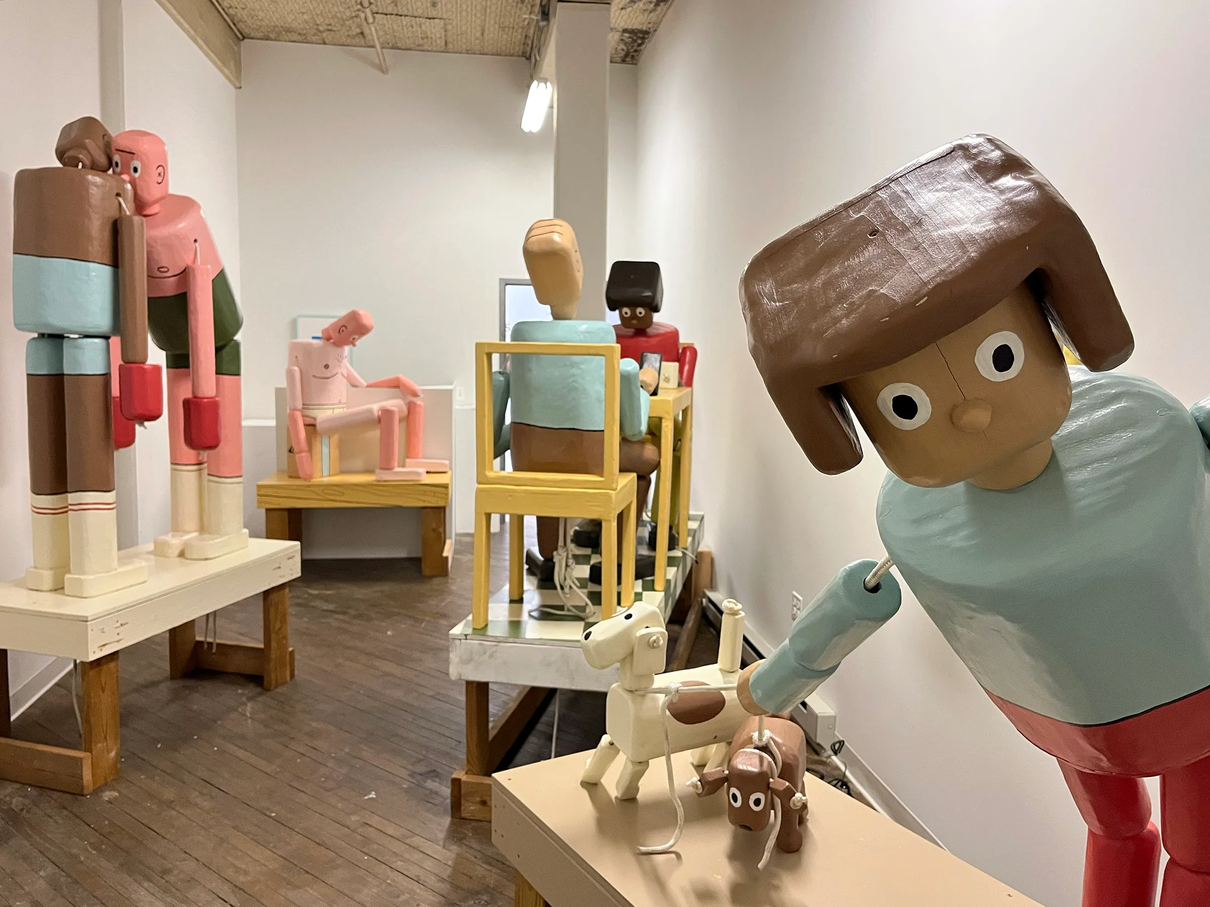 A photograph of an art installation featuring multiple figurative wooden sculptures. The life-sized sculptures resemble push puppet toys.