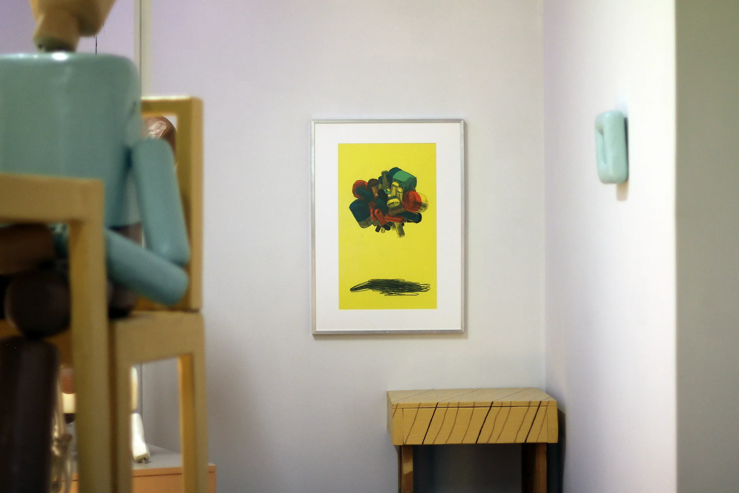 A photograph of a yellow, abstract drawing with a white mat in an aluminum frame. The frame hangs on a white gallery wall.