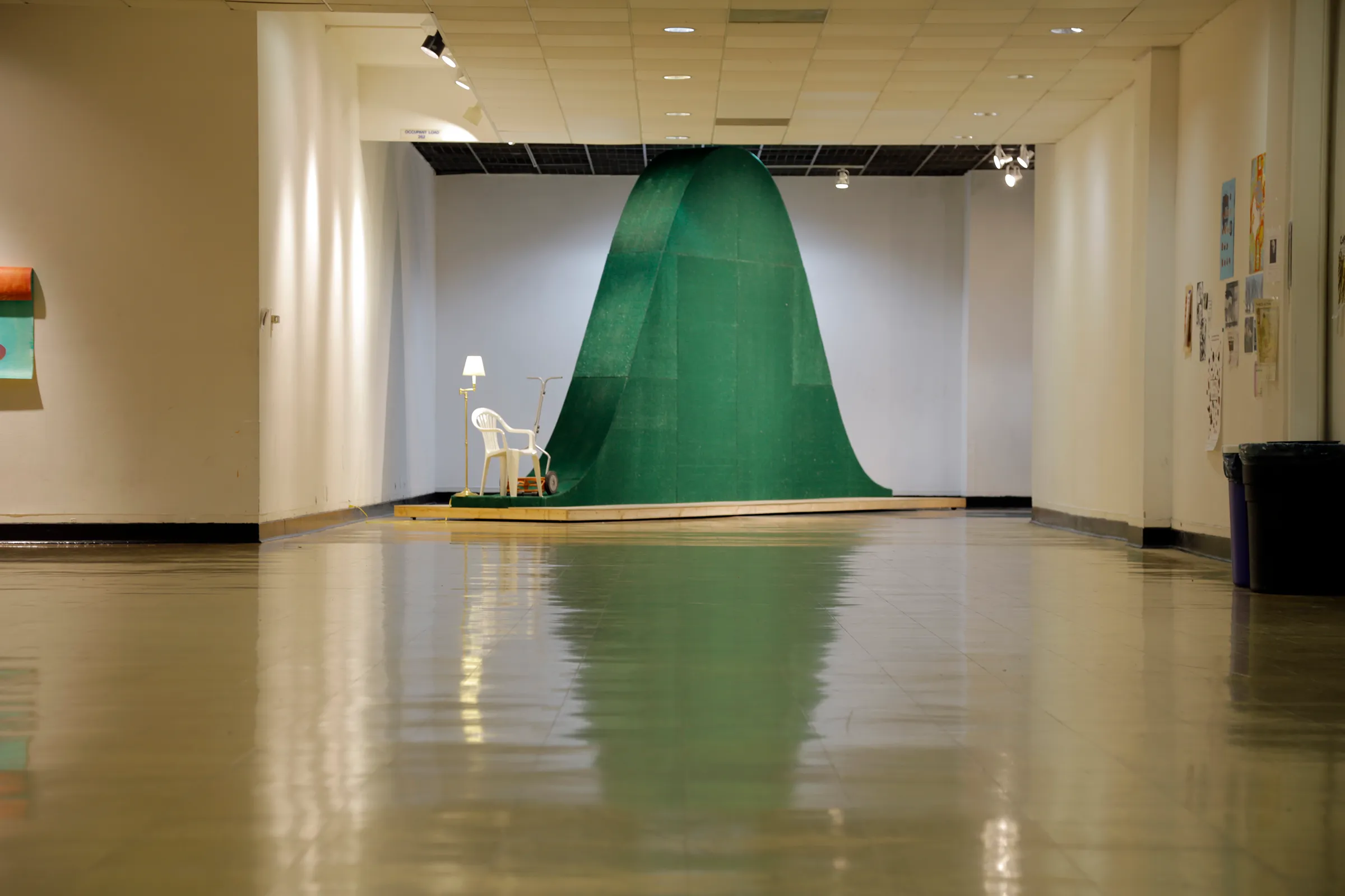 A photograph of a white plastic lawn chair next to a large, green, bell-curve shaped sculpture in a school hallway