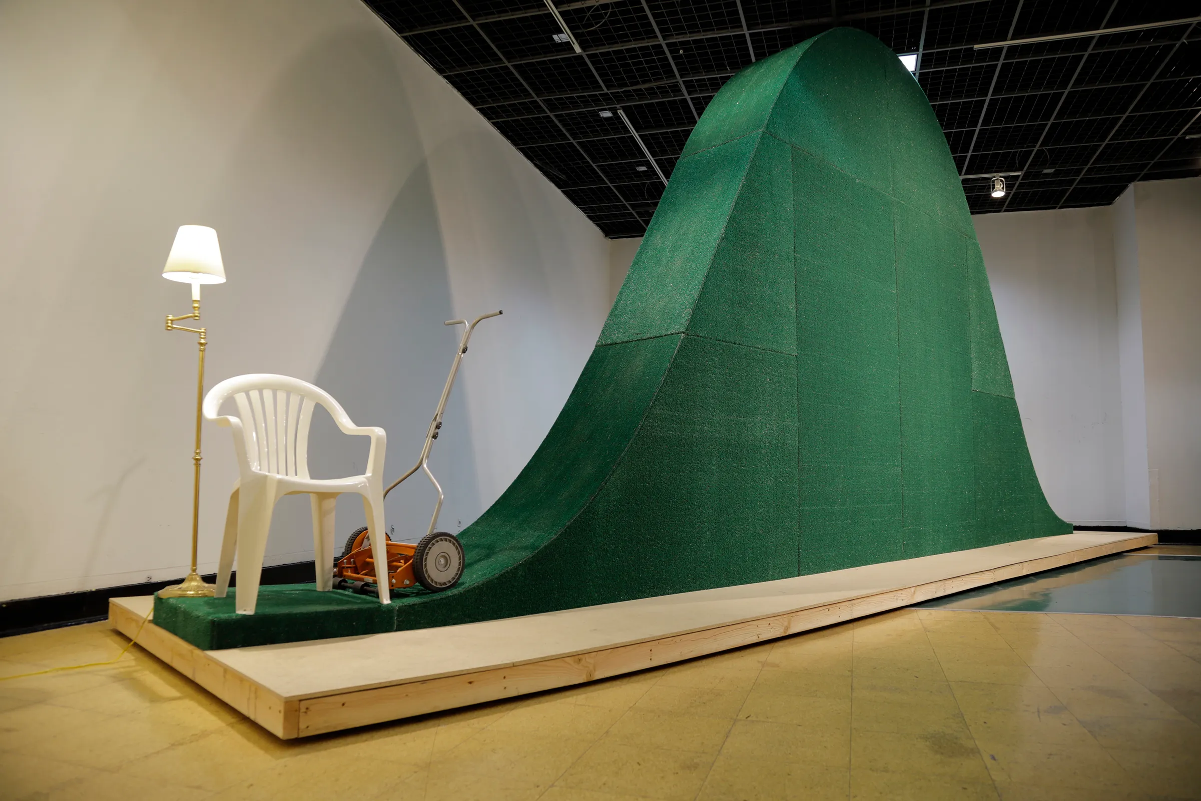 A close-up photograph of white plastic lawn chair, floor lamp, and push reel mower next to a large, green, bell-curve shaped sculpture.