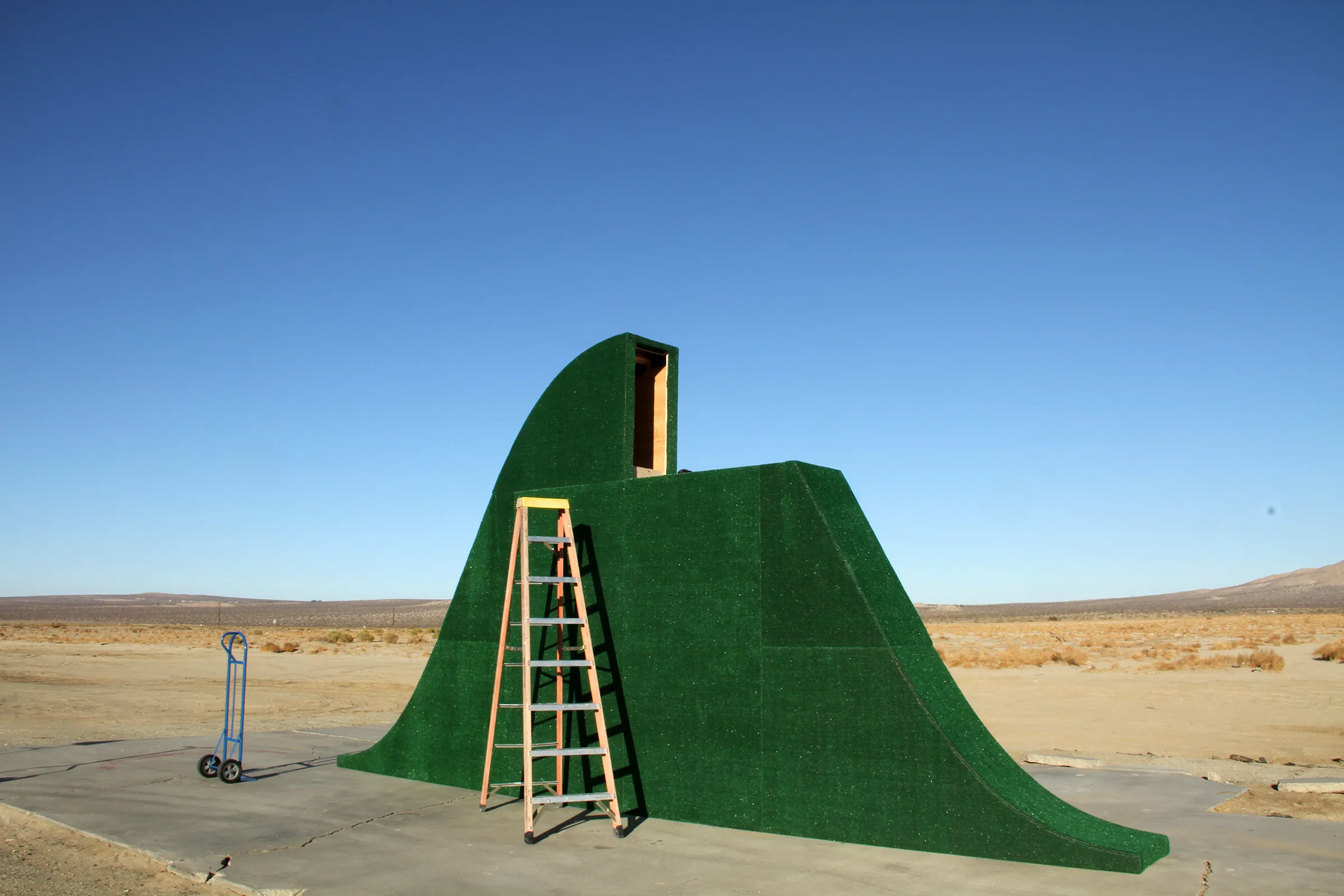 Two people assembling pieces of a large green sculpture in the middle of desert shrubland.