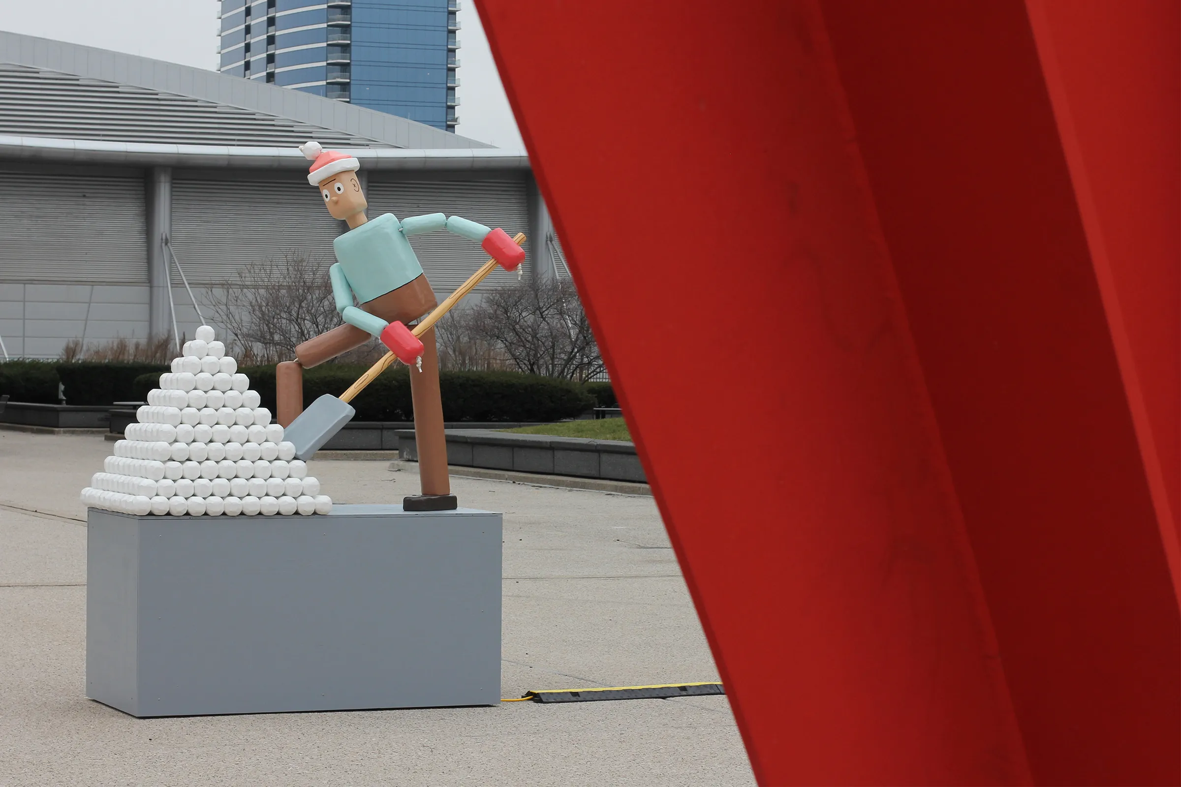 a bifurcated photograph of a snow-shoveling life-size push puppet sculpture made of wood on one side and a portion of a red metal sculpture on the other.