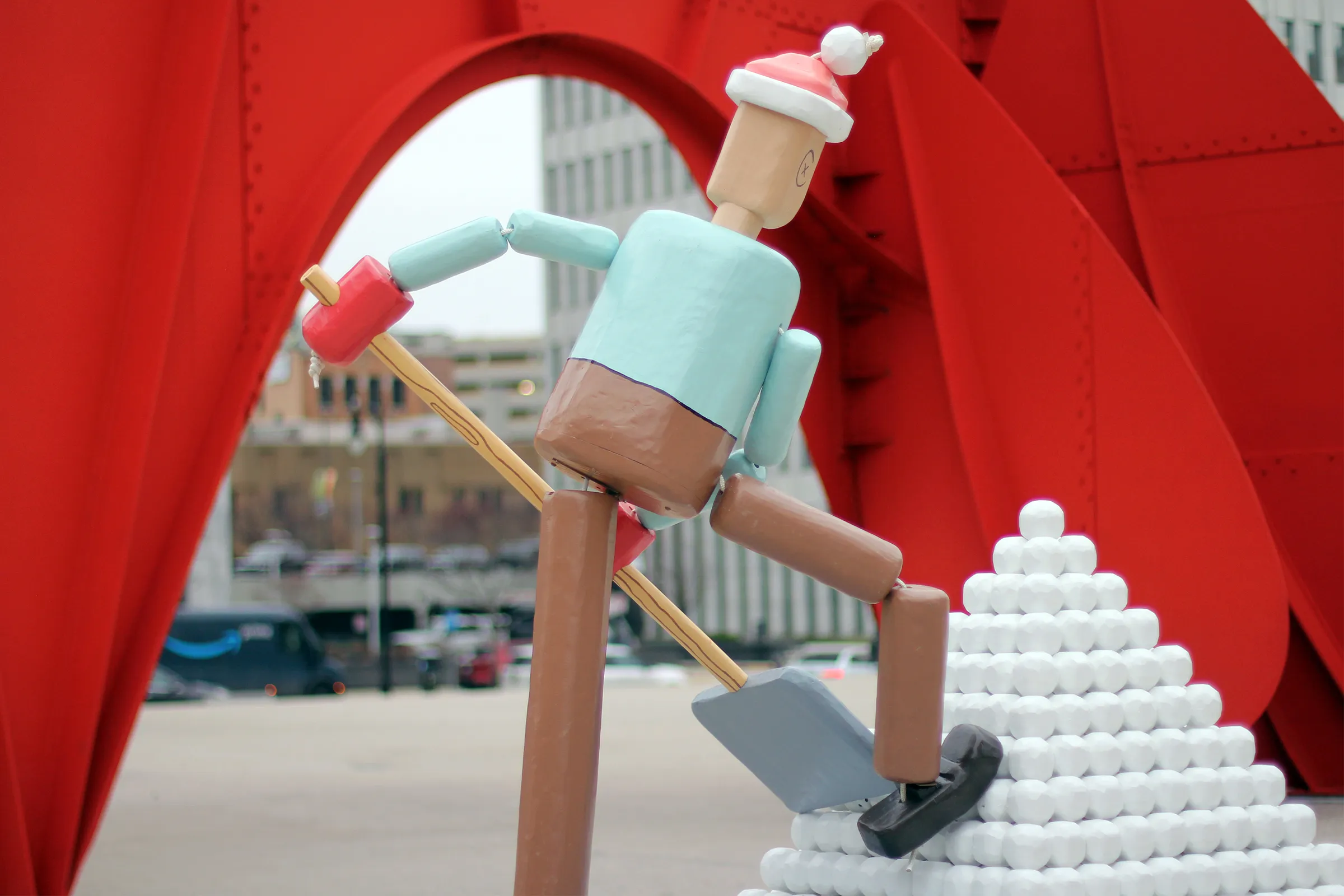 a rear view of a portion of a life-size push puppet sculpture in front a large, red metal sculpture in the background.