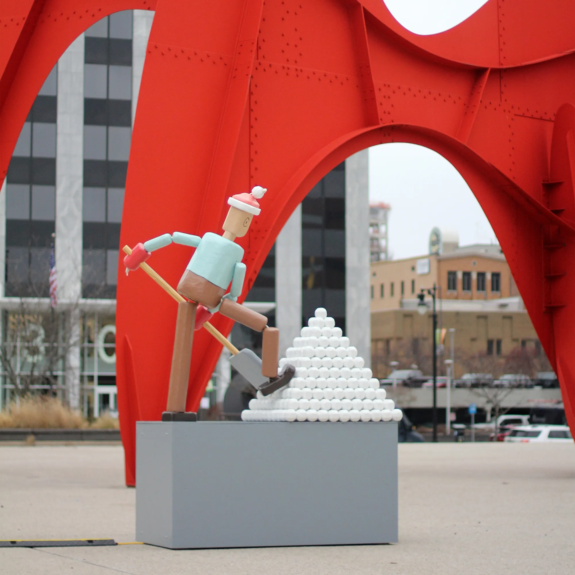 a rear view of the entire push puppet sculpture seen in front of a large, metal, public artwork.