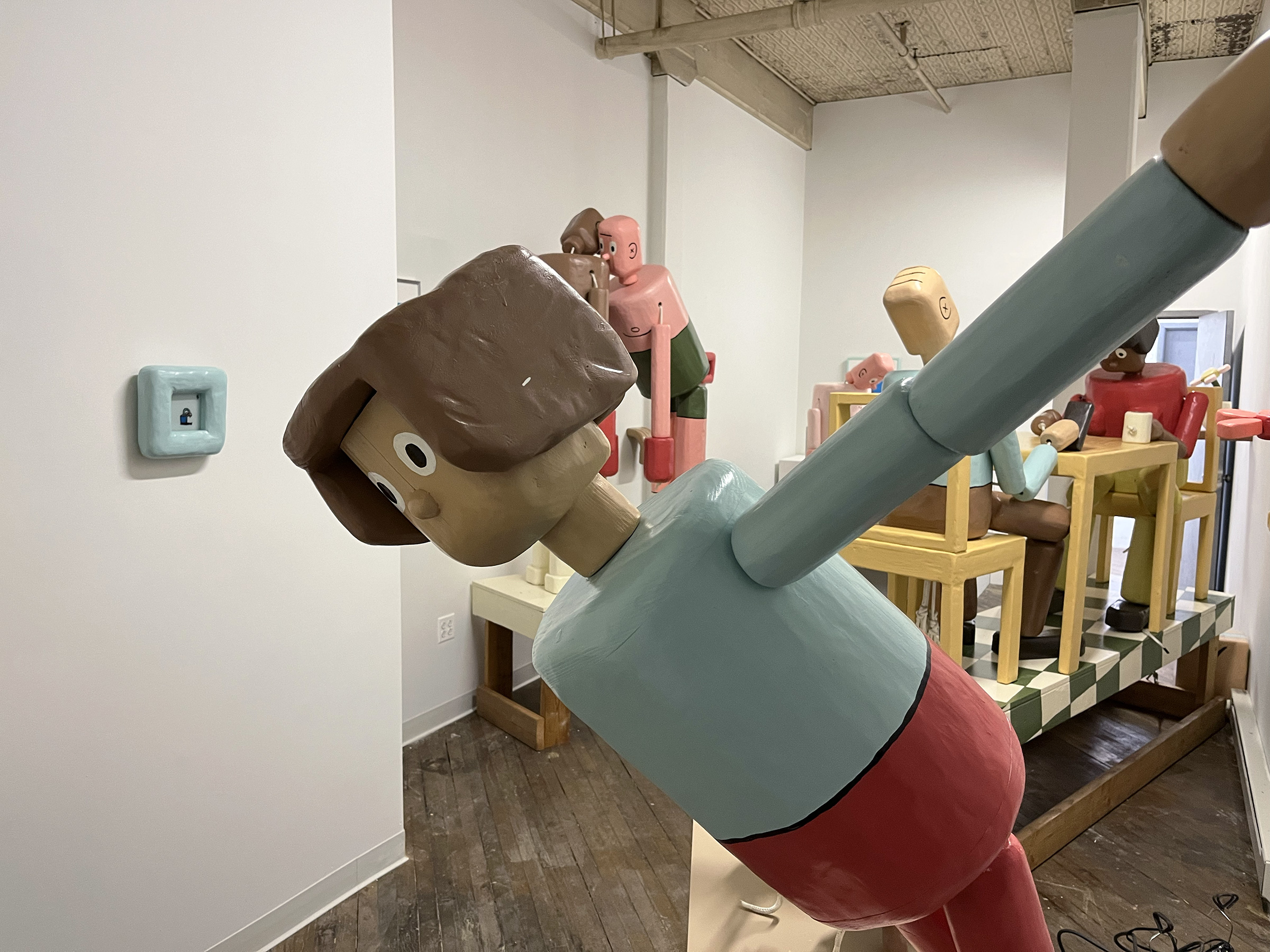 A photograph of an art installation featuring multiple figurative wooden sculptures. The life-sized sculptures resemble push puppet toys.