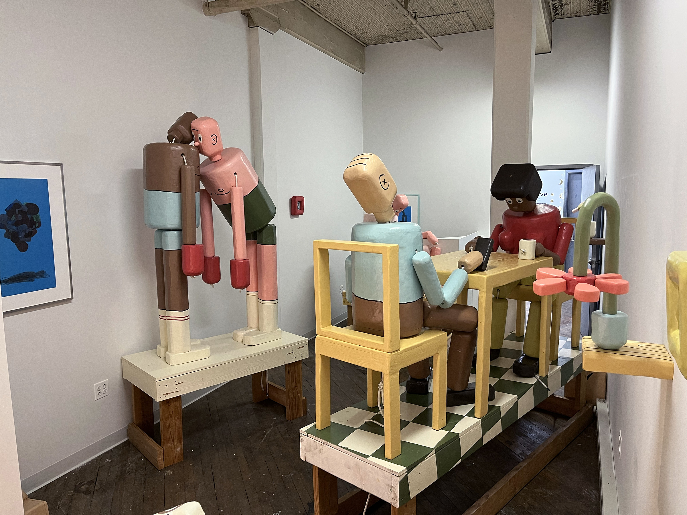 A photograph of an art installation featuring multiple figurative wooden sculptures. The life-sized sculptures resemble push puppet toys.