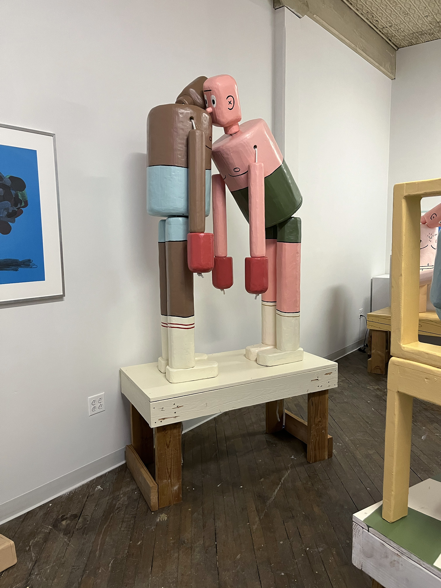A photograph of two wooden, life-sized, figurative sculptures that resemble push puppet toys. The two figures are painted to look like boxers. One sculpture’s head rests on the other sculpture’s shoulder.