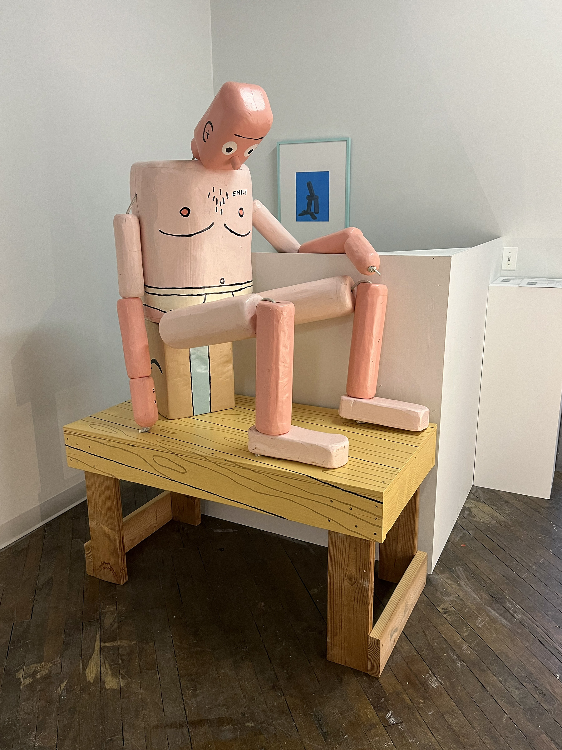 A photograph of a life-sized, figurative sculpture that resembles a push puppet toy. The wooden figure is painted to look like a man with pink skin wearing nothing but white underwear.