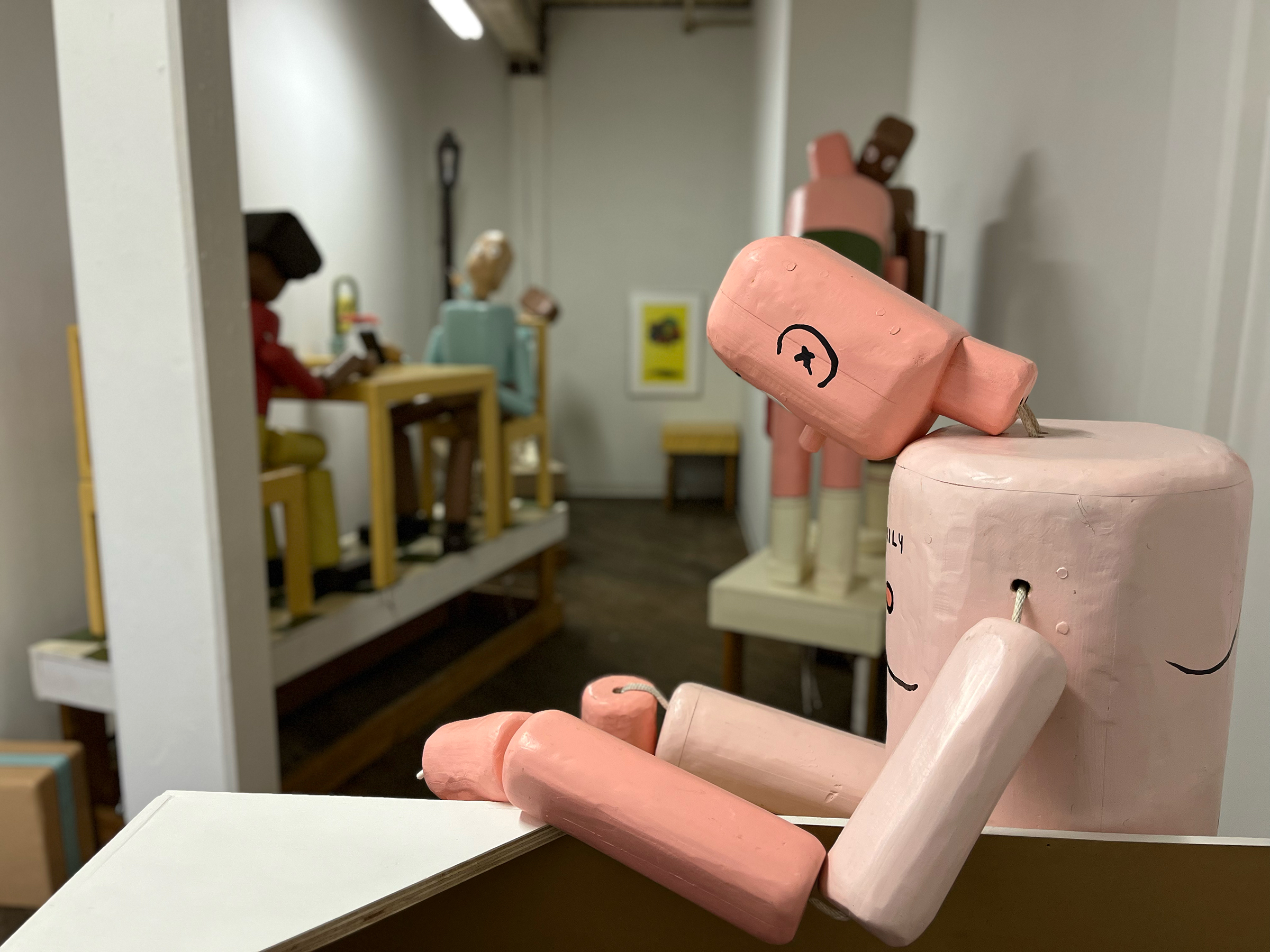 A photograph of a life-sized wooden sculpture of a man that resembles a push puppet toy in a gallery space with similar sculptures.