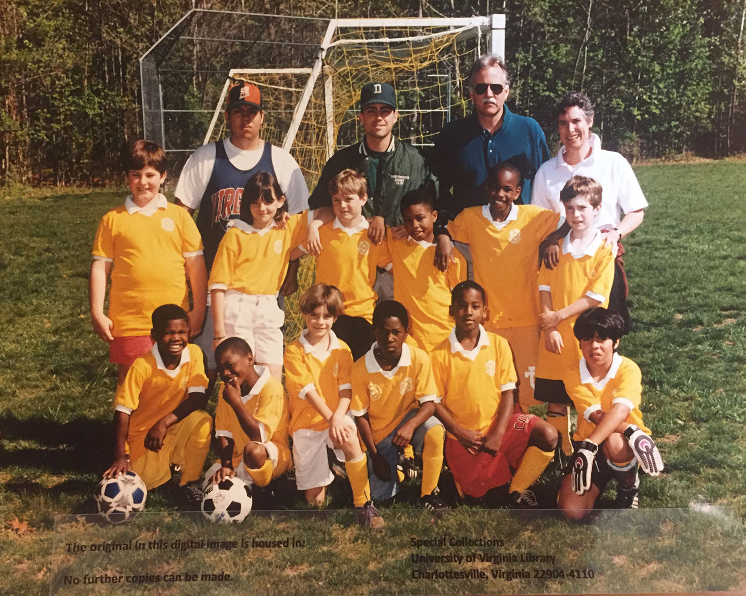 A photo of a youth soccer team from the 1990s. Wearing yellow jerseys, the team is arranged in two rows. The kids in the back stand and the kids in the front kneel. Four adult coaches stand behind the kids.