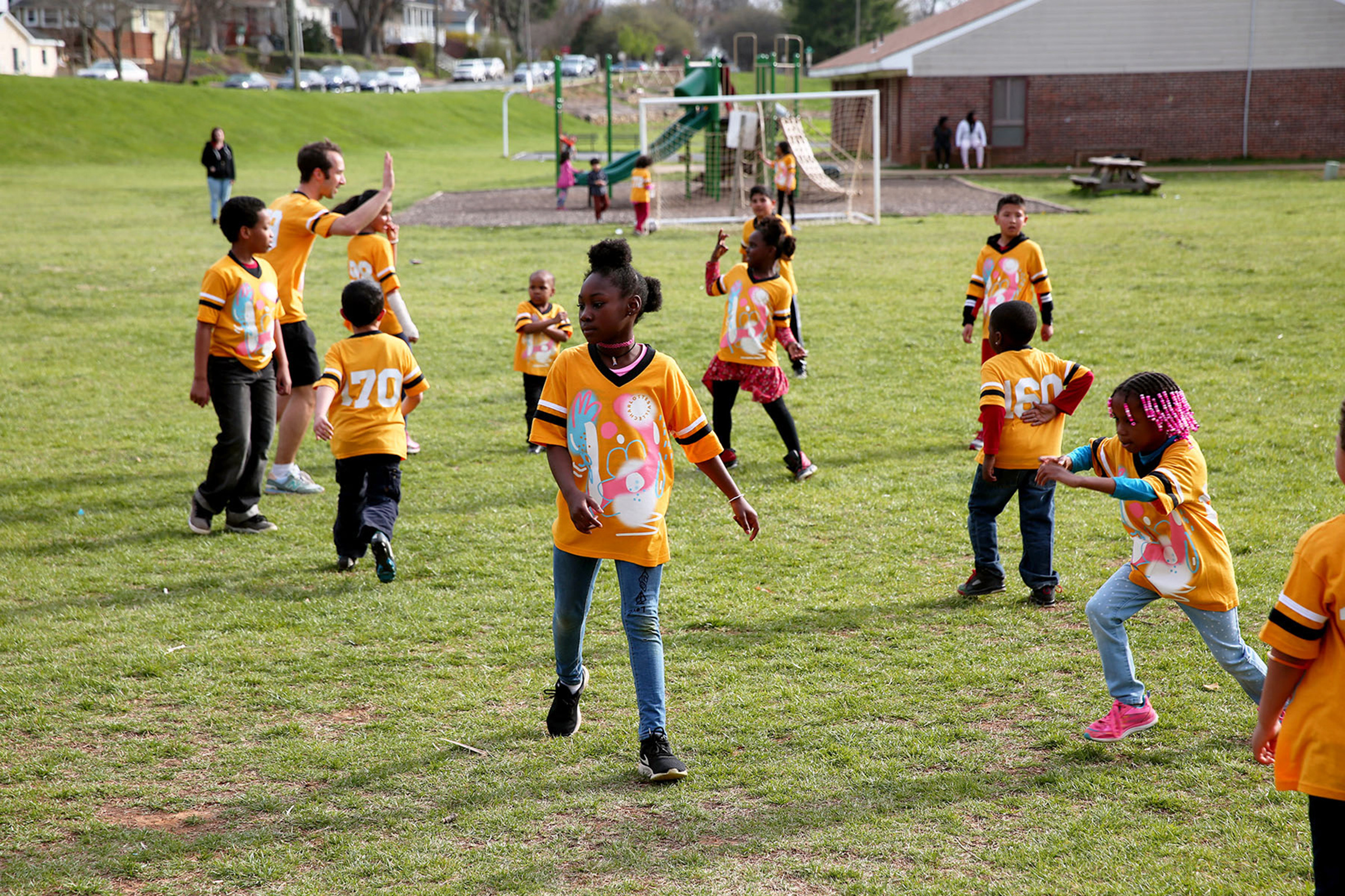 A dozen children and one adult wear matching yellow shirts while playing on a soccer field.