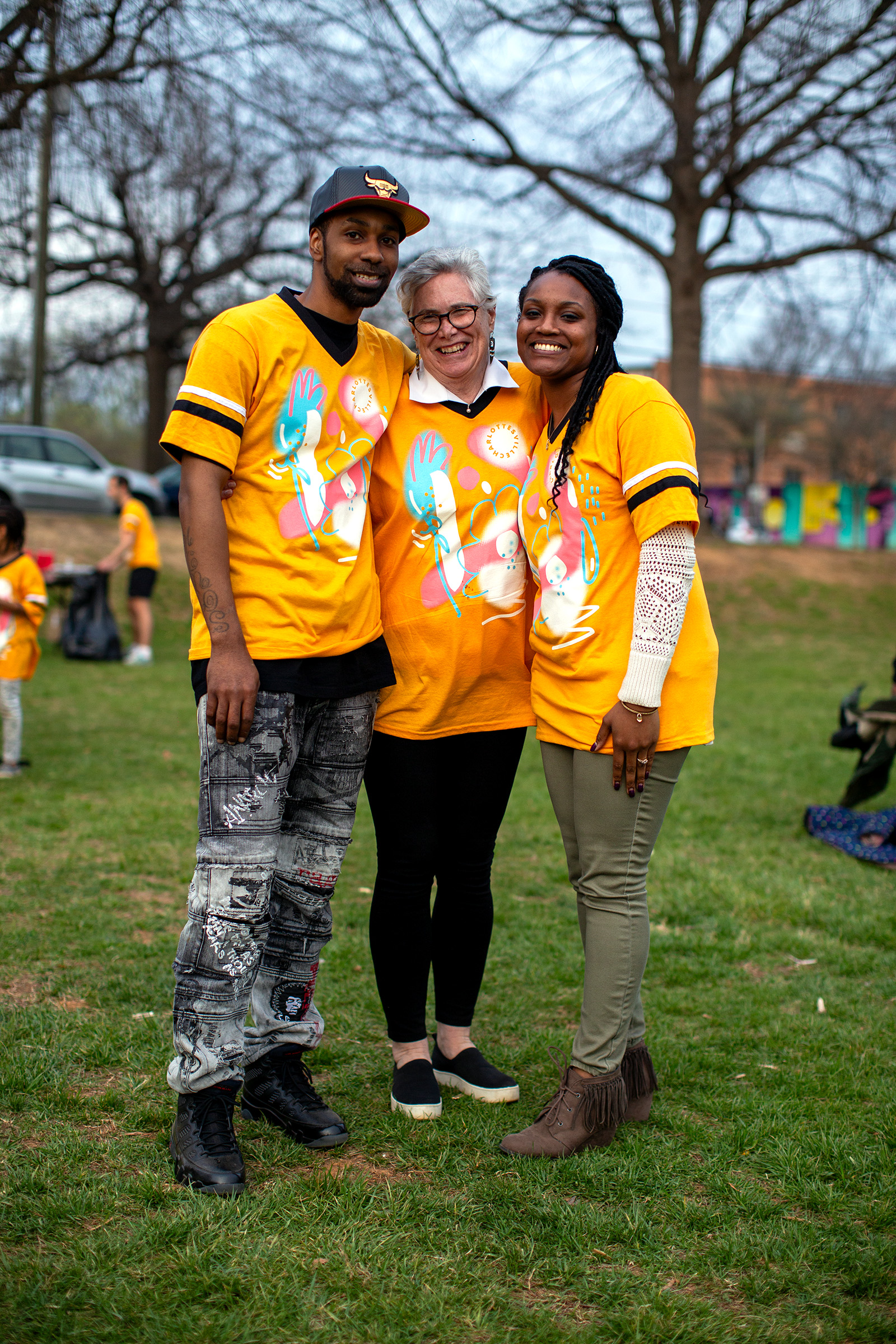 A photograph of three adults standing in a field wearing matching yellow jerseys that have an abstract floral graphic printed on them. The people have their arms around each other as they pose for the photo.
