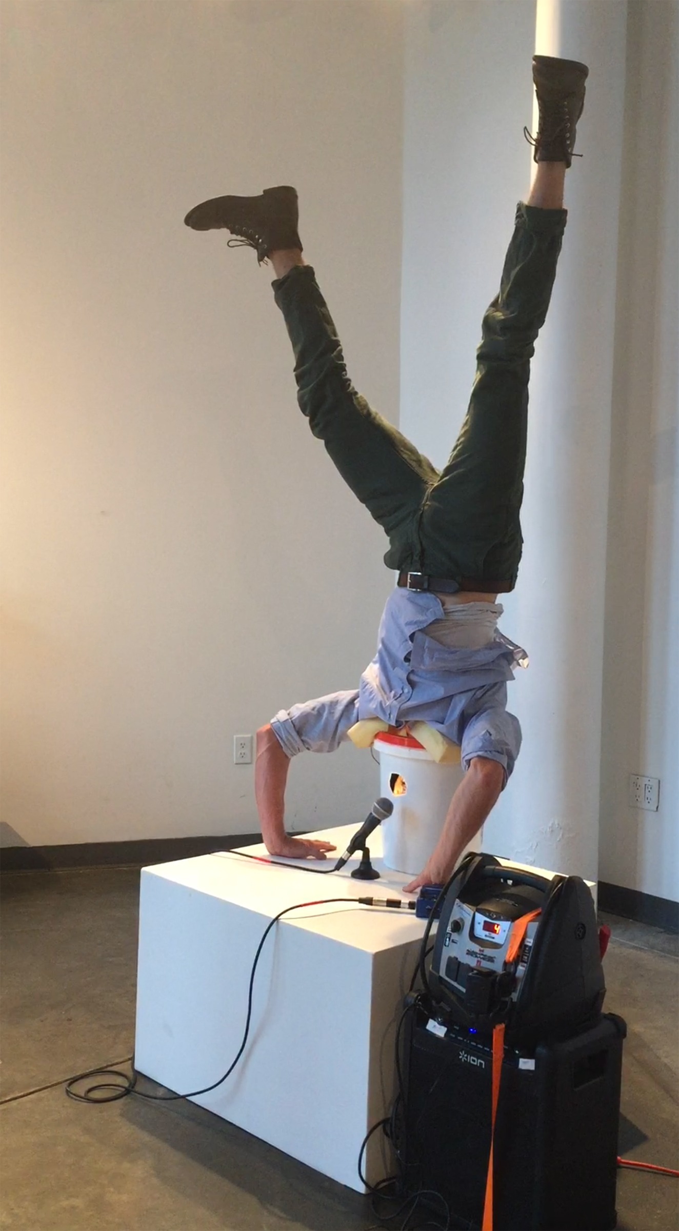 A photograph of Avery Lawrence doing a handstand with his head in a white 5-gallon bucket. He wears a light blue Oxford shirt, green pants, and brown leather boots.