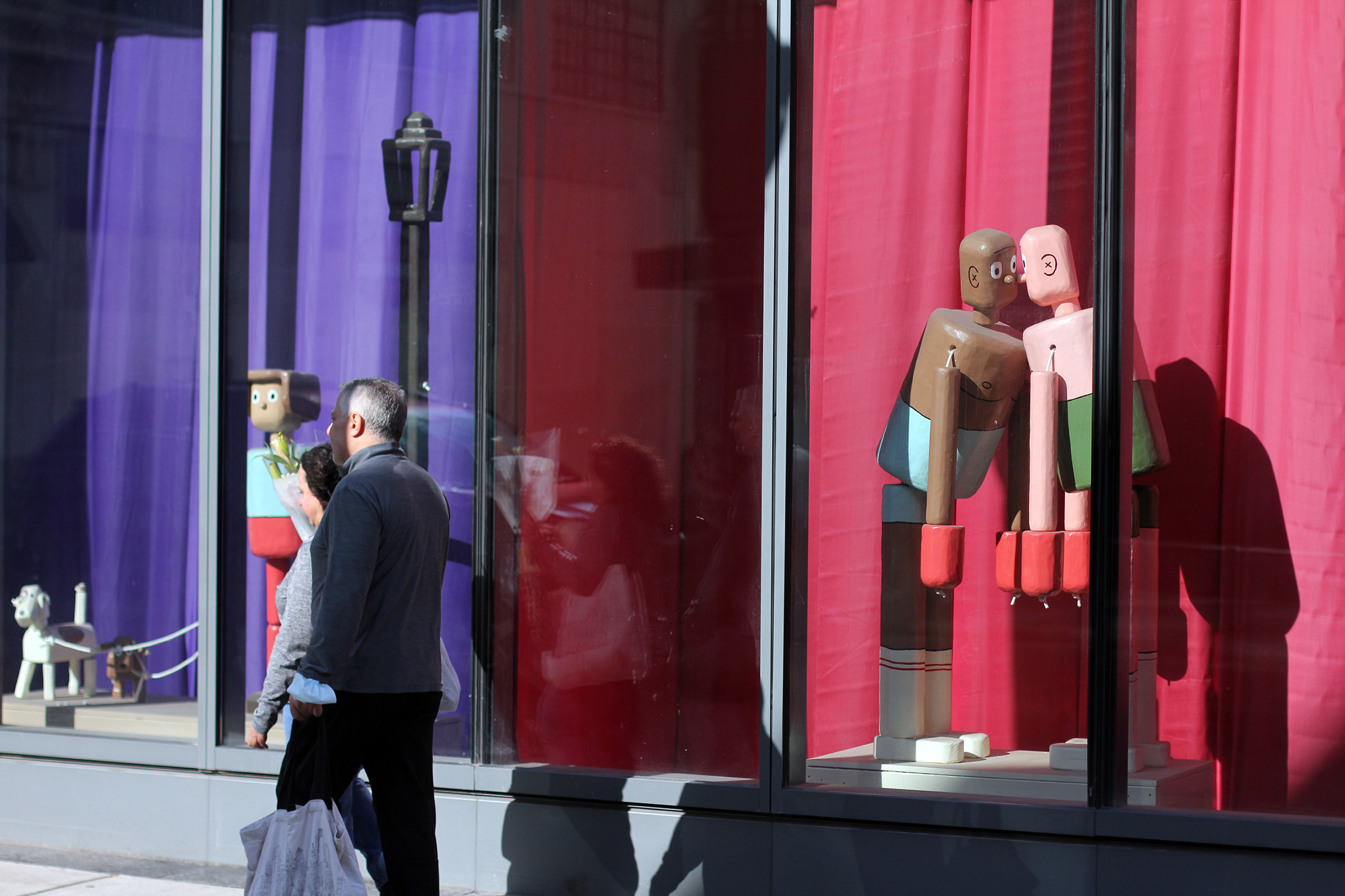 A photograph of two people walking past a window display featuring multiple life-size, wooden sculptures that look like push puppet toys.