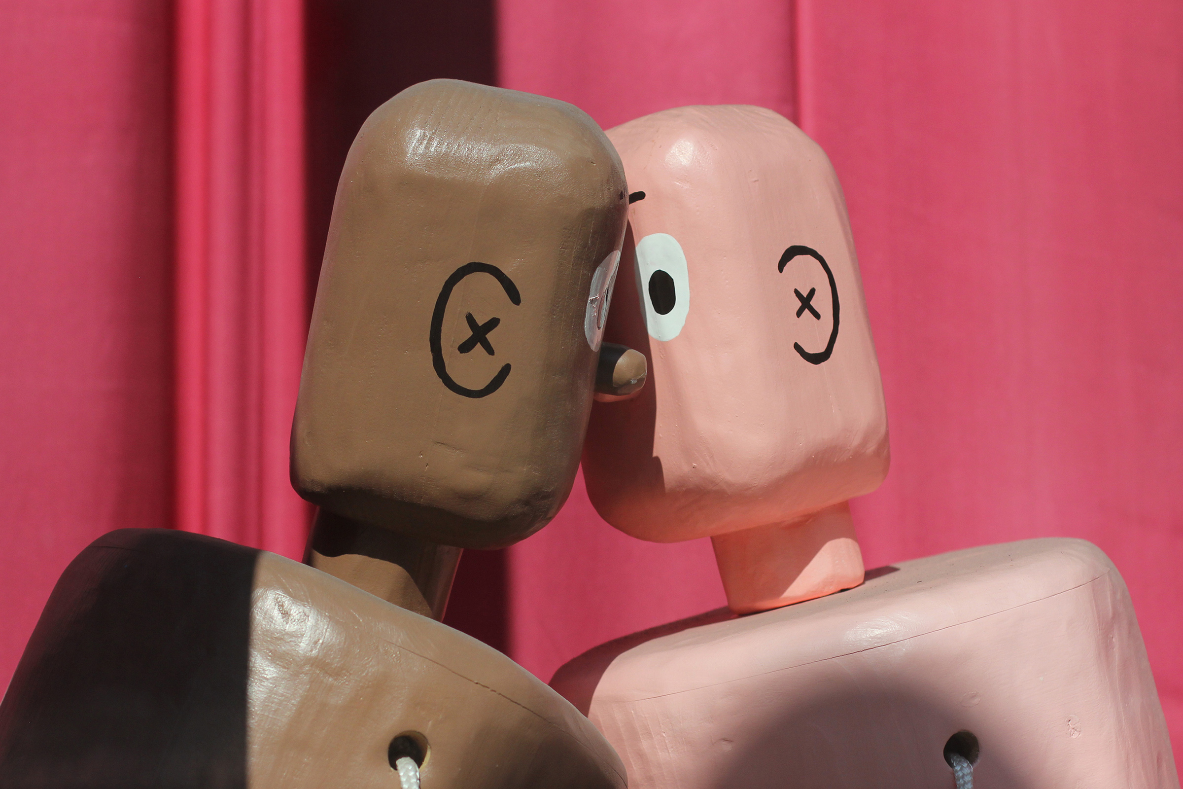 A closeup photograph of the heads and necks of two life-size, wooden sculptures painted to look like boxers that are leaning into each other. The figures resemble push puppet toys.