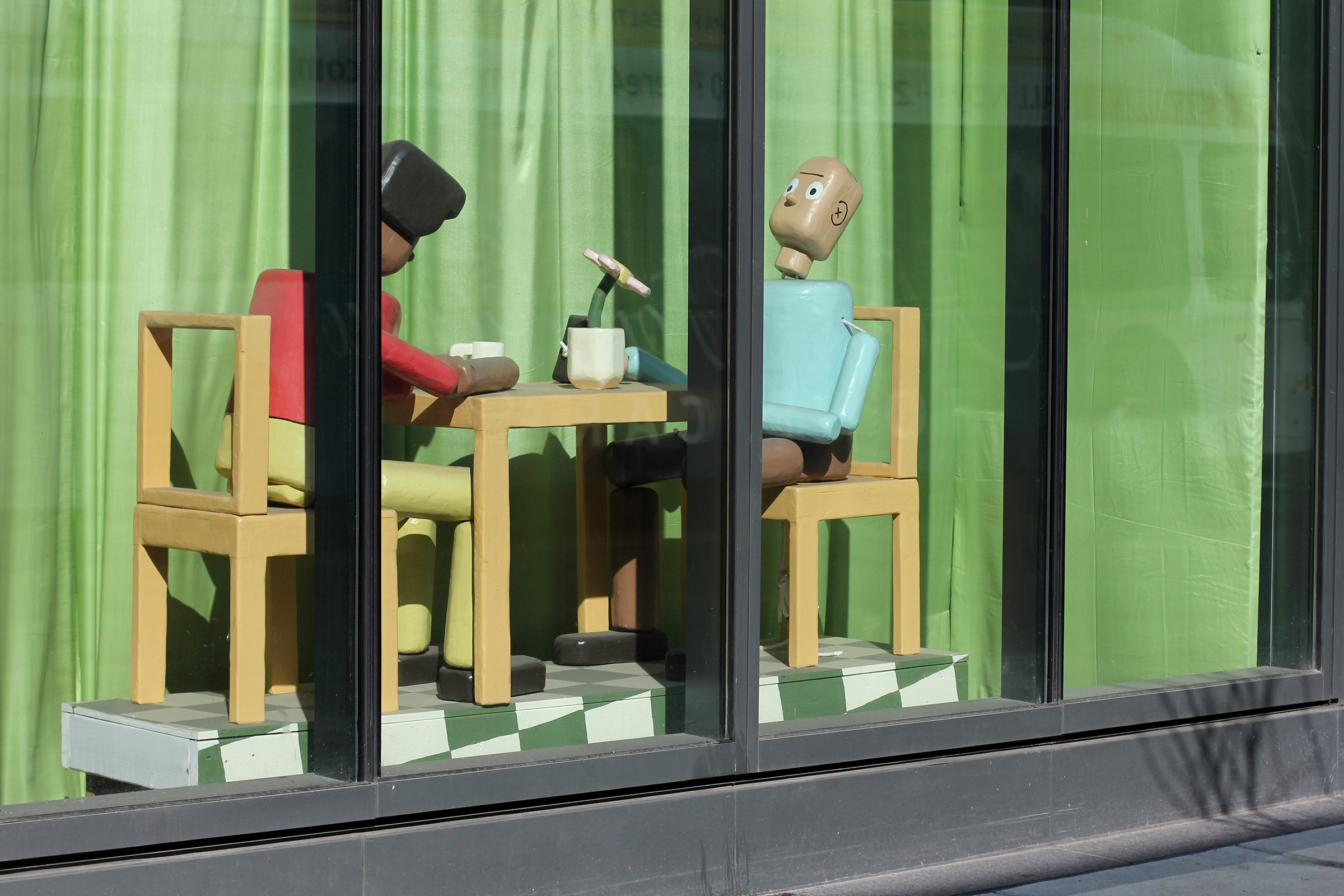 A photograph of two life-size, wooden sculptures painted to look like people sitting at a table, drinking coffee and looking at a phone. The figures resemble push puppet toys.