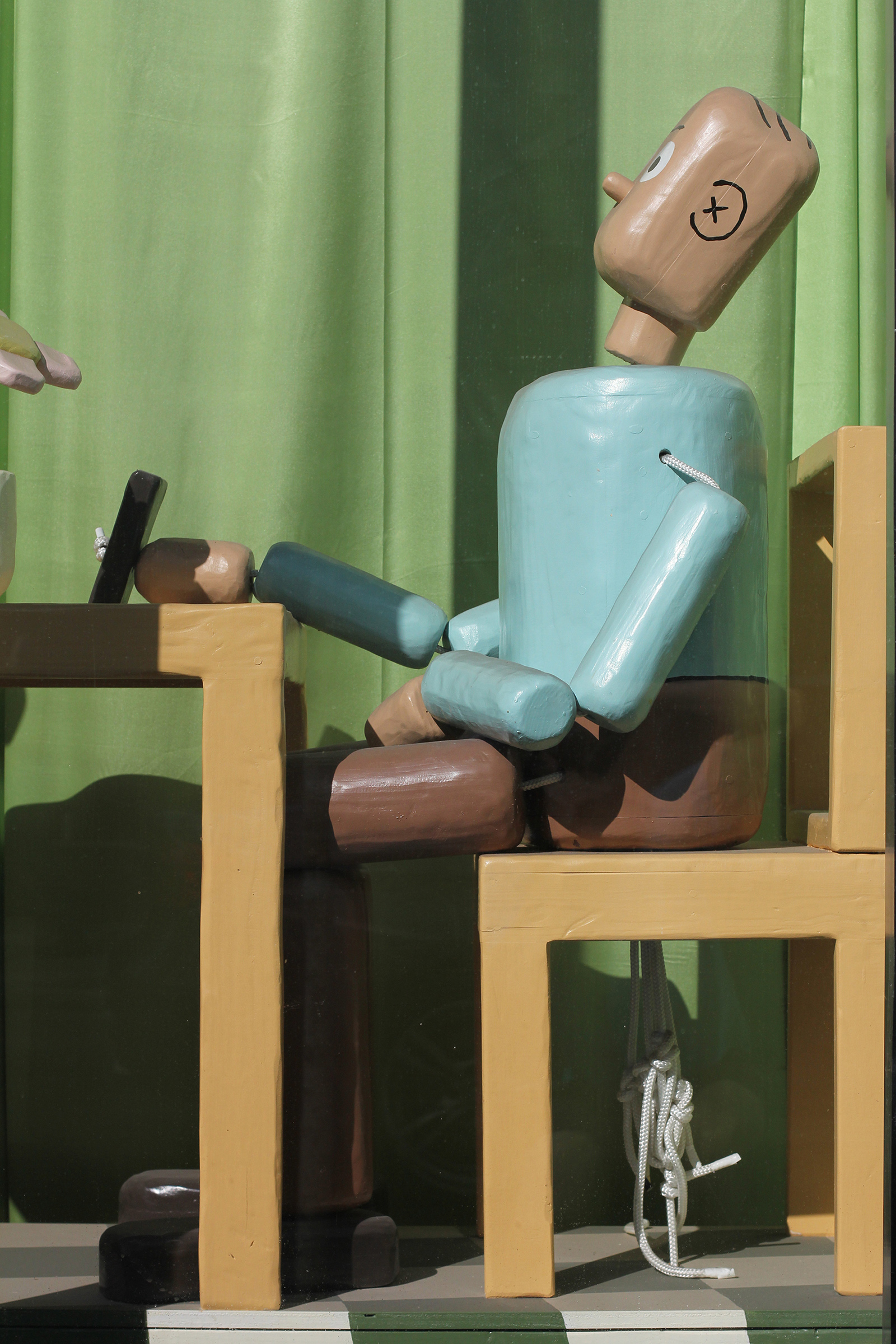 A closeup photograph of the wooden sculpture painted to look like a person sitting at a kitchen table. The figure, painted to have pink skin, resembles a push puppet toy.