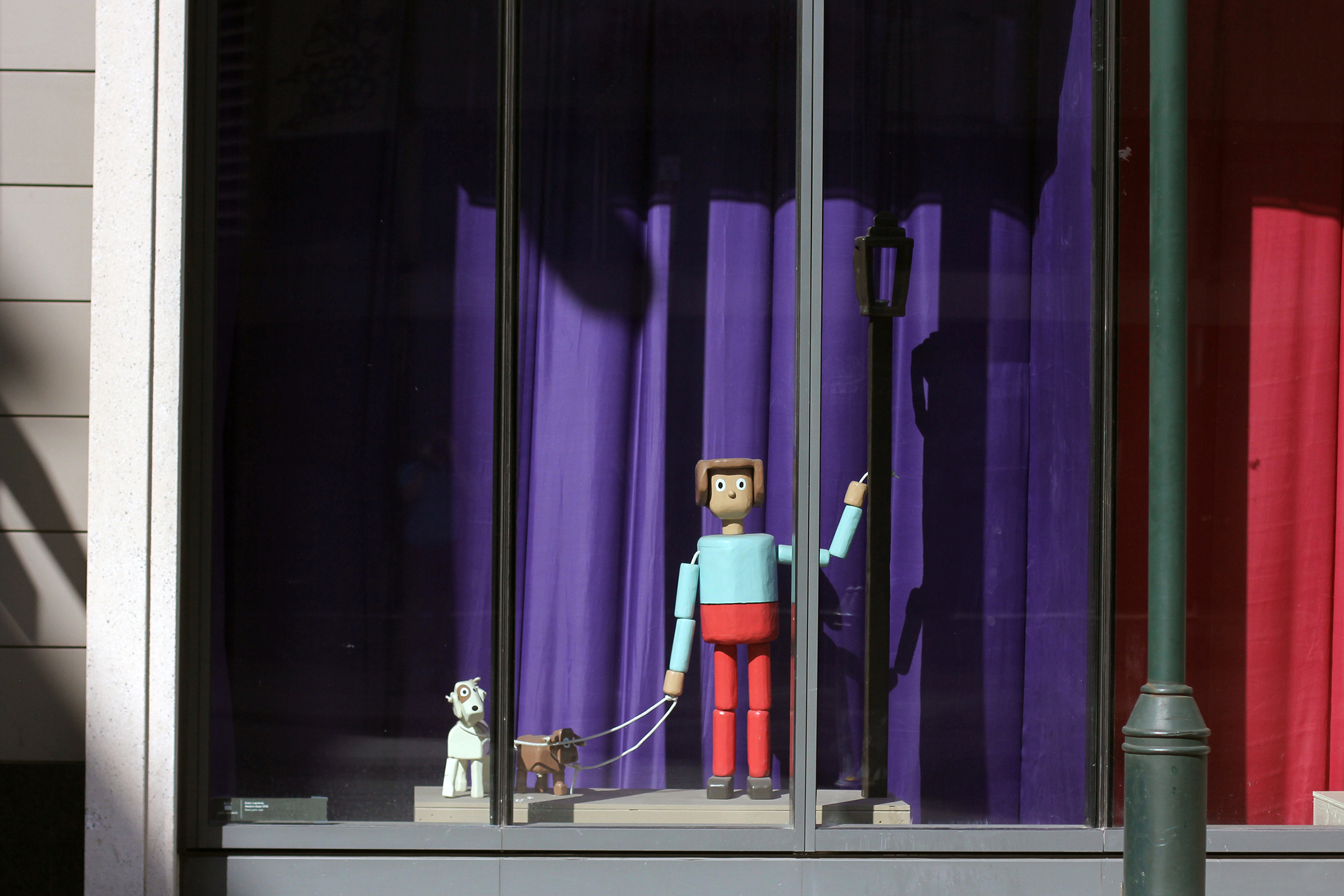 A photograph of a life-size, wooden sculpture painted to look like a person walking two dogs in a window display. The person and the dogs resemble push puppet toys.