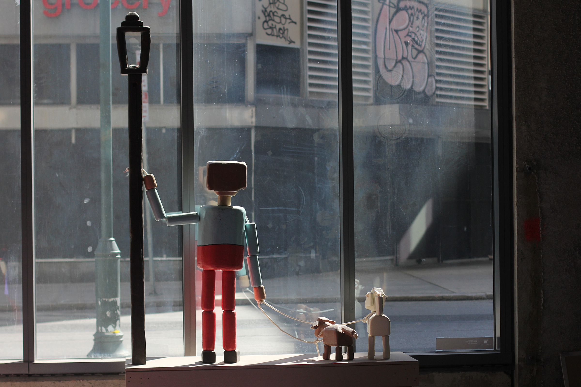 A photograph from the backside of a life-size, wooden sculpture painted to look like a person walking two dogs in a window display. The person and the dogs resemble push puppet toys.